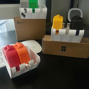Op de afbeelding: Een verzameling kleurrijke 3D-geprinte accessoires, waaronder grijze, groene, gele, zwarte, rode en oranje items, gepresenteerd in witte containers. De containers staan op bruine kartonnen dozen, met een witte Apple muis en toetsenbord.