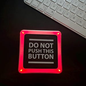 May include: A square, black button with the text "DO NOT PUSH THIS BUTTON" in white, surrounded by a glowing red frame. The button is on a dark surface, with a white keyboard in the background.