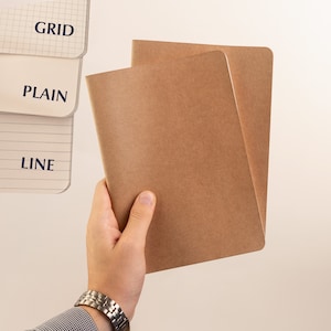 May include: Two brown kraft paper notebooks with blank covers. The notebooks are held in a hand.
