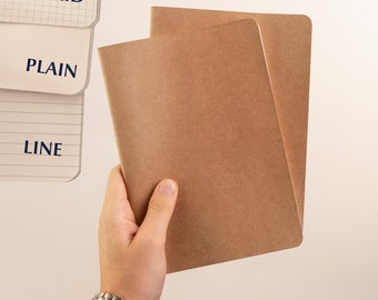 May include: Two brown kraft paper notebooks with blank covers. The notebooks are held in a hand.