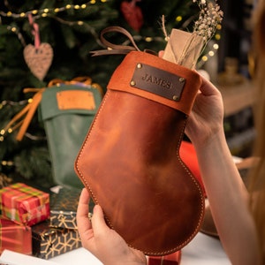 May include: A brown leather Christmas stocking with a suede top and a leather tag that reads "JAMES". The stocking is filled with gifts and dried flowers. The stocking is being held in front of a Christmas tree.