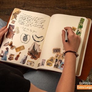 Pu&ograve; includere: Un diario aperto color rame extra large con copertina in pelle, pieno di adesivi decorativi e testo scritto a mano. Gli adesivi includono farfalle, disegni architettonici e illustrazioni botaniche. Una persona sta scrivendo nel diario con una penna.