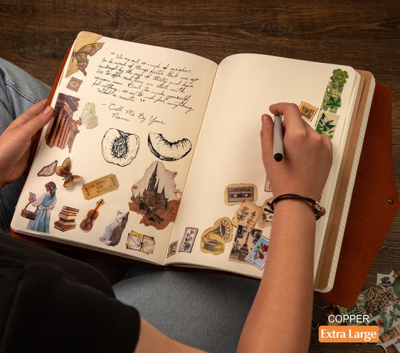 Pu&ograve; includere: Un diario aperto color rame extra large con copertina in pelle, pieno di adesivi decorativi e testo scritto a mano. Gli adesivi includono farfalle, disegni architettonici e illustrazioni botaniche. Una persona sta scrivendo nel diario con una penna.