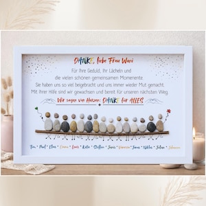 May include: A framed art print with a white border. The artwork features a message in German, with a pebble art design of people. The text includes names and a thank you message. A candle and vase are in the background.