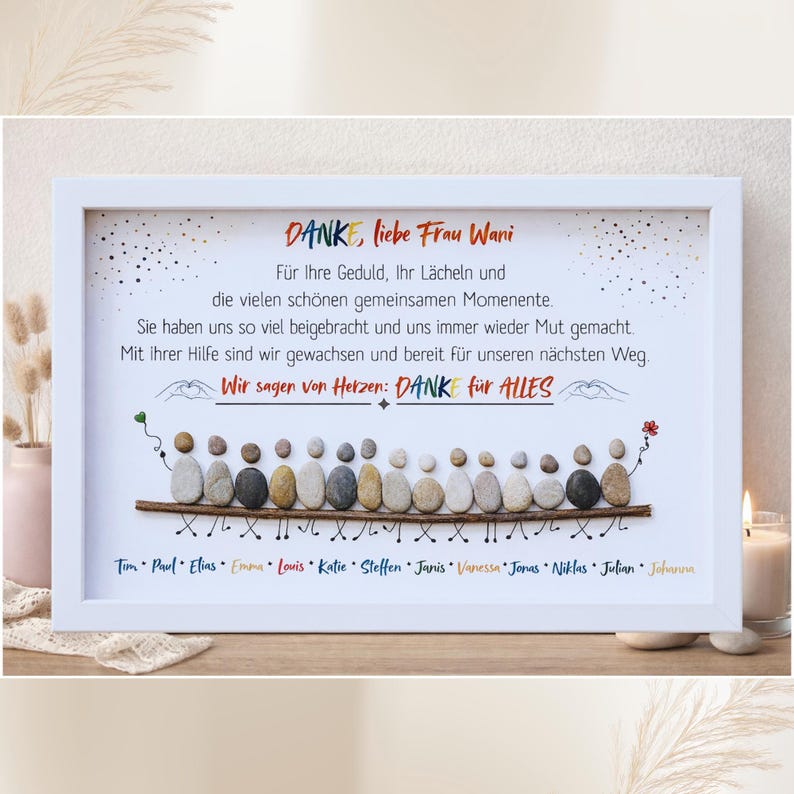 May include: A framed art print with a white border. The artwork features a message in German, with a pebble art design of people. The text includes names and a thank you message. A candle and vase are in the background.