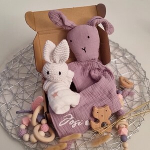 May include: A gift box filled with baby items, including a purple bunny plush toy, a white crocheted bunny toy, a wooden cat toy, and a wooden heart-shaped tag. The box is decorated with the name "Josie".
