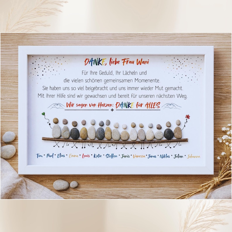 May include: A framed artwork with a white border. The artwork features a pebble art design of people and text in German. The text reads "DANK, liebe Frau Wani" and a message of gratitude. The names of the people are listed below the pebble art.