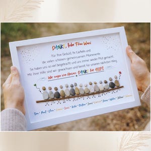 May include: A framed artwork with a white border, featuring a pebble art design. The artwork includes text in German, with a message of gratitude and a line of pebble figures. The names of individuals are listed below.