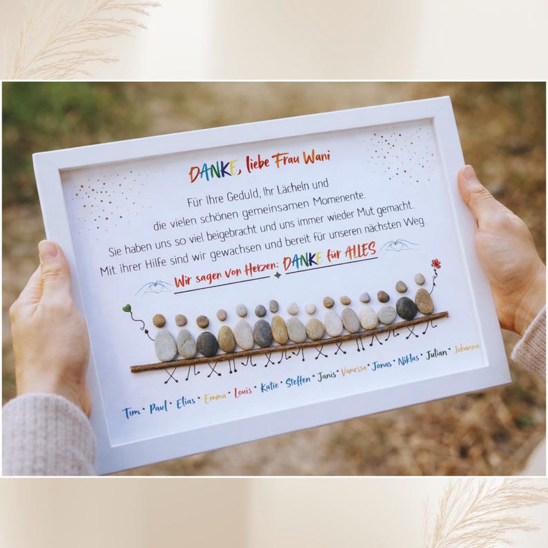 May include: A framed artwork with a white border, featuring a pebble art design. The artwork includes text in German, with a message of gratitude and a line of pebble figures. The names of individuals are listed below.