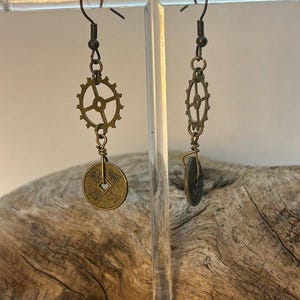 Puede incluir: Un par de pendientes de estilo steampunk. Un pendiente presenta un engranaje de color bronce y una moneda, el otro un engranaje y una piedra oscura y redonda. Los pendientes cuelgan de ganchos para las orejas de color bronce.