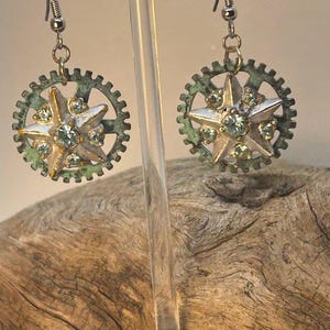 Puede incluir: Un par de pendientes de estilo steampunk. Cada pendiente presenta un colgante en forma de estrella con piedras preciosas transparentes, engastado en un marco en forma de engranaje con pátina verde. Los pendientes cuelgan de ganchos plateados.