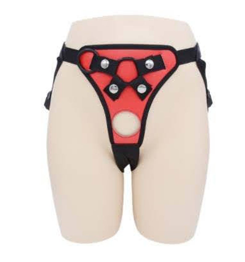 May include: A red and black leather harness with adjustable straps and metal buckles. The harness has a large, circular opening in the front.