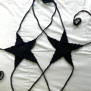 May include: Black crochet top featuring two star-shaped cups connected by straps. The top is handmade and appears to be made of a soft yarn. The star shapes have pointed edges and are designed to be worn as a halter top.