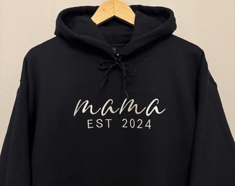 Könnte beinhalten: Schwarzer Kapuzenpullover mit dem Wort "mama" in weißer Schrift und "EST 2024" darunter gedruckt. Der Kapuzenpullover hat einen Kordelzug und eine Kapuze. Das Kleidungsstück hängt an einem Holzbügel.