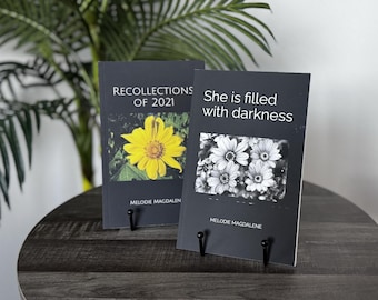 May include: Two black paperbacks with white text on the covers. The book on the left has a yellow flower on the cover and the text "Recollections of 2021" and "Melodie Magdalen". The book on the right has a black and white flower on the cover and the text "She is filled with darkness" and "Melodie Magdalene".