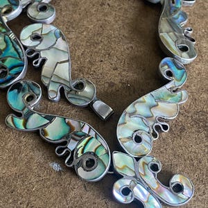 Peut inclure: Un collier compos&eacute; de segments m&eacute;talliques argent&eacute;s reli&eacute;s entre eux et incrust&eacute;s de morceaux de nacre iris&eacute;e. Le design pr&eacute;sente des formes courbes en volutes avec une finition polie, affichant un spectre de tons bleus, verts et nacr&eacute;s.