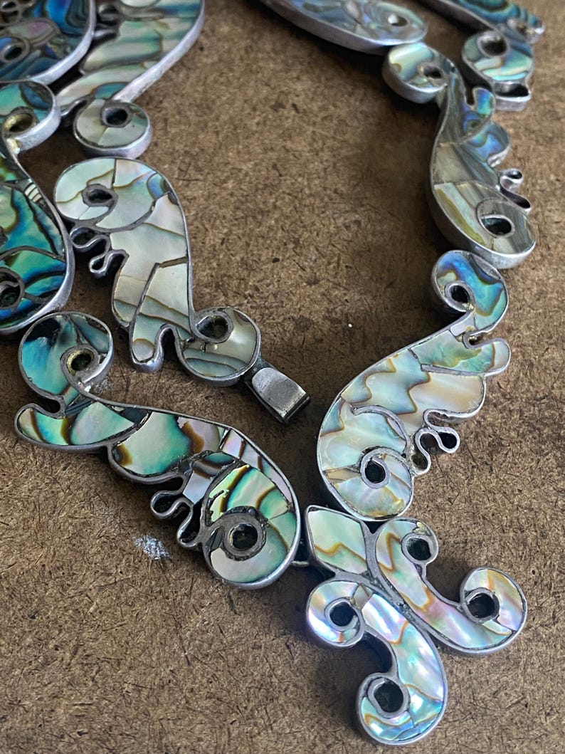 Peut inclure: Un collier compos&eacute; de segments m&eacute;talliques argent&eacute;s reli&eacute;s entre eux et incrust&eacute;s de morceaux de nacre iris&eacute;e. Le design pr&eacute;sente des formes courbes en volutes avec une finition polie, affichant un spectre de tons bleus, verts et nacr&eacute;s.