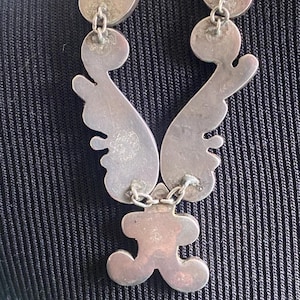 Peut inclure: Un collier en m&eacute;tal argent&eacute; compos&eacute; de plusieurs segments interconnect&eacute;s aux formes courbes et organiques, reli&eacute;s par de petits anneaux m&eacute;talliques. La surface du m&eacute;tal pr&eacute;sente une finition mate et l&eacute;g&egrave;rement patin&eacute;e.
