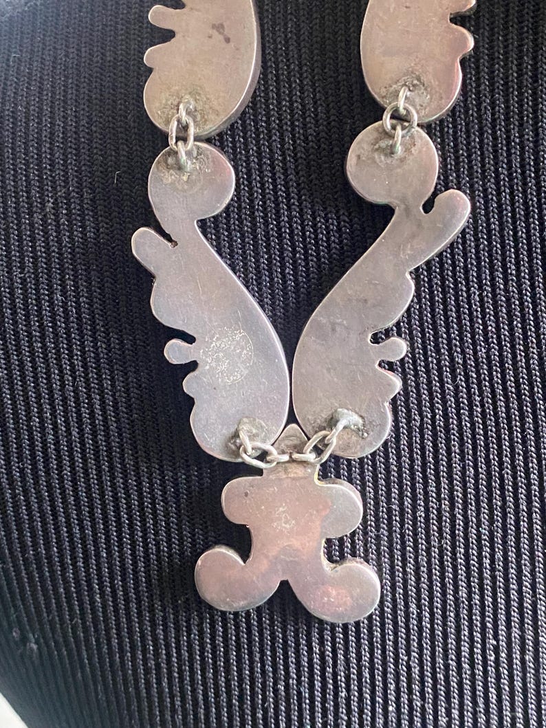 Peut inclure: Un collier en m&eacute;tal argent&eacute; compos&eacute; de plusieurs segments interconnect&eacute;s aux formes courbes et organiques, reli&eacute;s par de petits anneaux m&eacute;talliques. La surface du m&eacute;tal pr&eacute;sente une finition mate et l&eacute;g&egrave;rement patin&eacute;e.