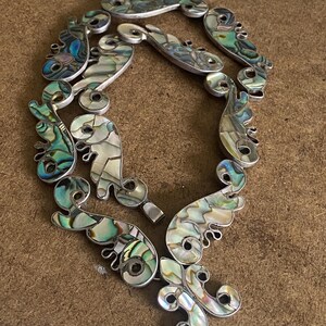 Peut inclure: Un collier compos&eacute; de multiples segments reli&eacute;s entre eux, incrust&eacute;s de morceaux de nacre d'ormeau iris&eacute;e. Les segments pr&eacute;sentent des formes organiques incurv&eacute;es avec des d&eacute;tails en spirale, affichant un spectre de tons bleus, verts et blancs nacr&eacute;s.