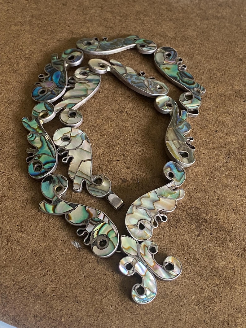 Peut inclure: Un collier compos&eacute; de multiples segments reli&eacute;s entre eux, incrust&eacute;s de morceaux de nacre d'ormeau iris&eacute;e. Les segments pr&eacute;sentent des formes organiques incurv&eacute;es avec des d&eacute;tails en spirale, affichant un spectre de tons bleus, verts et blancs nacr&eacute;s.