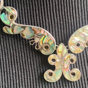 Peut inclure: Un collier aux tons argent&eacute;s compos&eacute; de maillons m&eacute;talliques incurv&eacute;s en forme de volutes, incrust&eacute;s de nacre d'ormeau iris&eacute;e. Le pendentif central est une fleur de lys stylis&eacute;e, &eacute;galement remplie de segments de nacre aux reflets changeants.