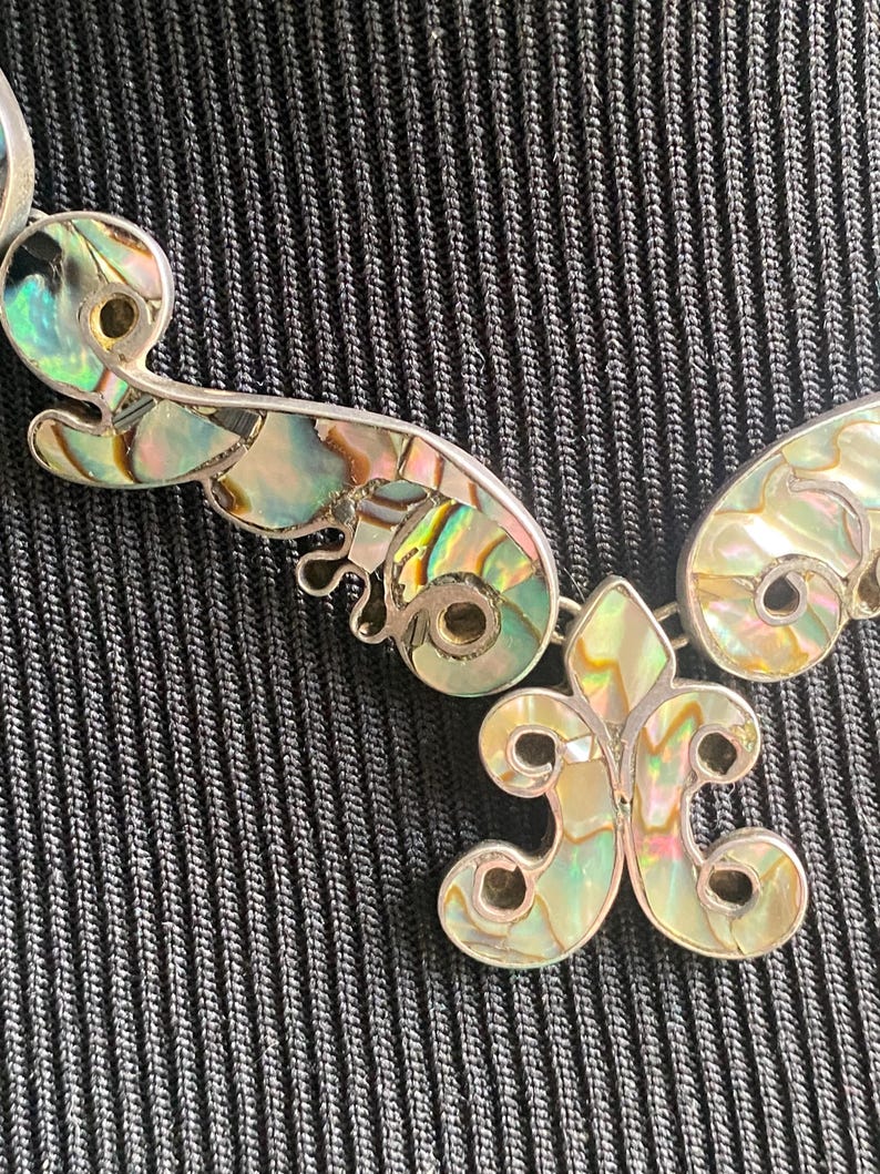 Peut inclure: Un collier aux tons argent&eacute;s compos&eacute; de maillons m&eacute;talliques incurv&eacute;s en forme de volutes, incrust&eacute;s de nacre d'ormeau iris&eacute;e. Le pendentif central est une fleur de lys stylis&eacute;e, &eacute;galement remplie de segments de nacre aux reflets changeants.