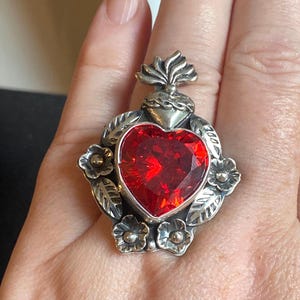 Op de afbeelding: Een zilveren ring met een grote, gefacetteerde, hartvormige rode steen in het midden. De zetting is versierd met reliëfmotieven van bloemen en bladeren, en een klein, gestileerd vlamelement zit boven op het hart voor een gedetailleerd ontwerp.