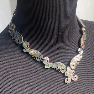 Peut inclure: Un collier argent&eacute; compos&eacute; d'une s&eacute;rie de maillons m&eacute;talliques incurv&eacute;s et interconnect&eacute;s, incrust&eacute;s de nacre d'ormeau iris&eacute;e. Le design comprend un pendentif central en forme de fleur de lys, chaque segment affichant des motifs tourbillonnants aux teintes vertes, bleues et nacr&eacute;es.