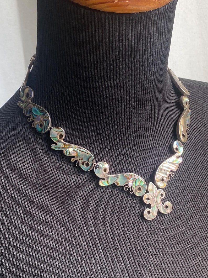 Peut inclure: Un collier argent&eacute; compos&eacute; d'une s&eacute;rie de maillons m&eacute;talliques incurv&eacute;s et interconnect&eacute;s, incrust&eacute;s de nacre d'ormeau iris&eacute;e. Le design comprend un pendentif central en forme de fleur de lys, chaque segment affichant des motifs tourbillonnants aux teintes vertes, bleues et nacr&eacute;es.