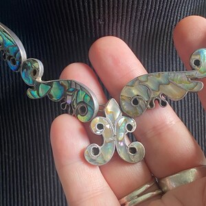 Peut inclure: Un collier argent&eacute; compos&eacute; de plusieurs segments articul&eacute;s incrust&eacute;s de nacre d'ormeau iris&eacute;e. Le pendentif central pr&eacute;sente une forme de fleur de lys, tandis que les maillons environnants arborent des motifs incurv&eacute;s en volutes avec des reflets bleus, verts et cr&egrave;me.