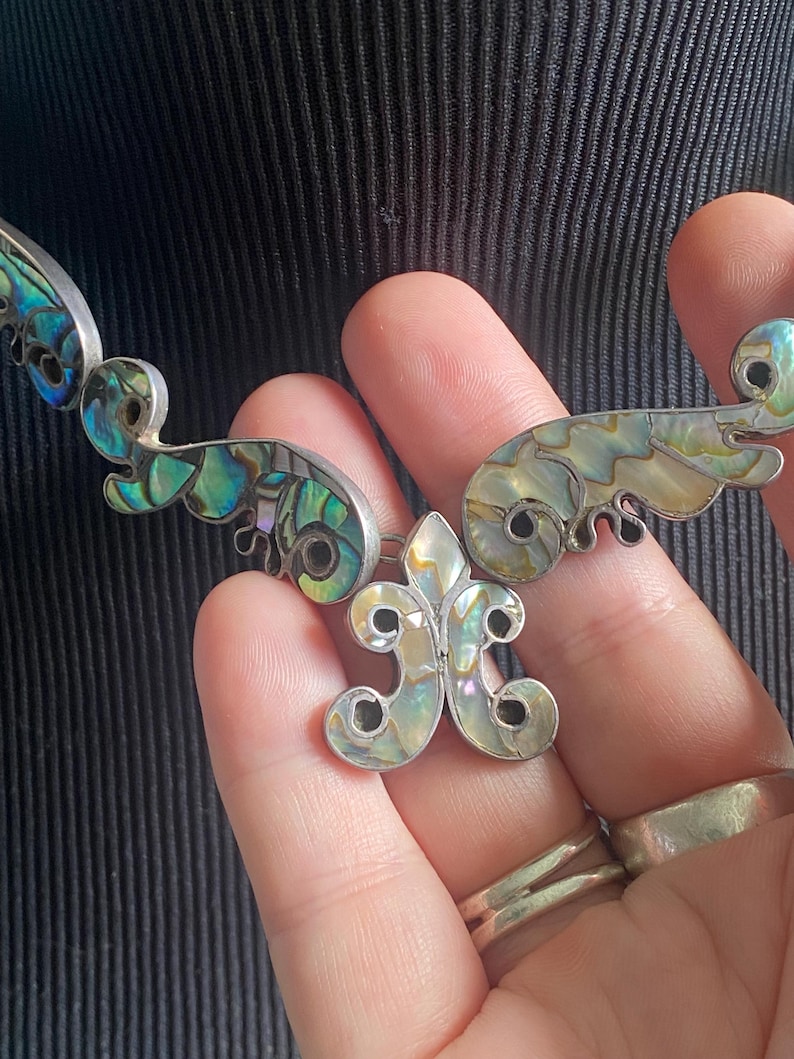 Peut inclure: Un collier argent&eacute; compos&eacute; de plusieurs segments articul&eacute;s incrust&eacute;s de nacre d'ormeau iris&eacute;e. Le pendentif central pr&eacute;sente une forme de fleur de lys, tandis que les maillons environnants arborent des motifs incurv&eacute;s en volutes avec des reflets bleus, verts et cr&egrave;me.