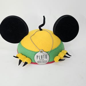 May include: A Pluto-themed hat with large black ears, a yellow top, and green and red trim. The hat has a black curly tail and yellow paws with black claws. A silver tag reads "PLUTO" and "if found, please return to MICKEY MOUSE."