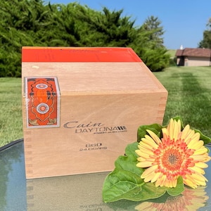Puede incluir: Una caja de cigarros de madera con el texto "Cain Daytona" y "660 24 Cigars" se muestra. La caja tiene una parte superior naranja y una etiqueta decorativa. Una margarita amarilla y naranja descansa junto a la caja.