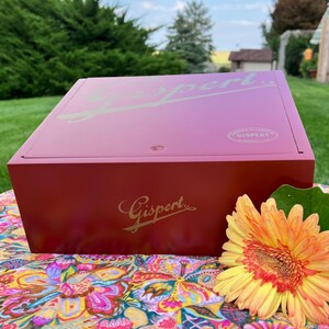 Puede incluir: Una caja de madera roja con una inscripción dorada que dice "Gispert" en la parte superior e inferior. La caja está sobre una tela floral colorida.