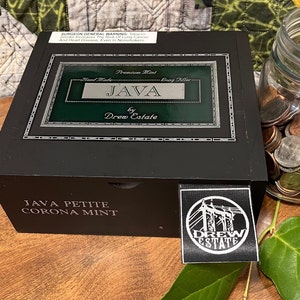 Op de afbeelding: Een zwarte houten doos met een groen label waarop staat "Premium Mint Hand Made - Bang Filler JAVA by Drew Estate". De doos is gelabeld "JAVA PETITE CORONA MINT" en heeft een zwart-witte sticker met de tekst "DREW ESTATE" en een afbeelding van een stroomlijn.
