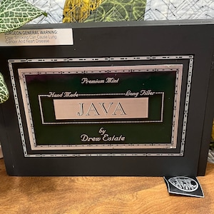 Op de afbeelding: Een zwarte houten doos met een groen en zilveren label waarop "Premium Mint Hand Made Long Filler JAVA by Drew Estate" staat.