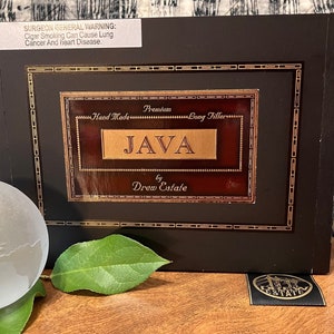 May include: A dark brown wooden box with a gold trim and a label that reads "Premium Hand Made Long Filler JAVA by Drew Estate". The box is sitting on a wooden surface with a green leaf and a clear glass globe.