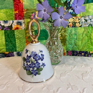 Pode incluir: Um sino de porcelana branca com uma alça dourada e um desenho floral de violetas roxas. O sino está sentado em uma superfície branca com um vaso de flores roxas no fundo.