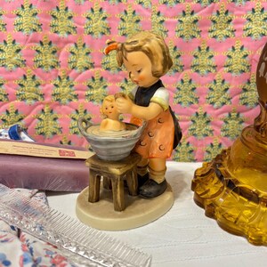 Puede incluir: Una figurita de porcelana de una niña con un vestido rojo y blanco bañando a una muñeca en una pequeña bañera. La niña sostiene una toallita y la muñeca está sentada en la bañera con agua jabonosa. La figurita está sobre un taburete de madera y tiene una base blanca.