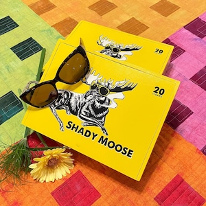 May include: Two yellow boxes of "Shady Moose" cigars, each containing 20 cigars, with a black and white moose graphic wearing sunglasses. Tortoiseshell sunglasses rest on top, with a yellow flower and greenery in the foreground.