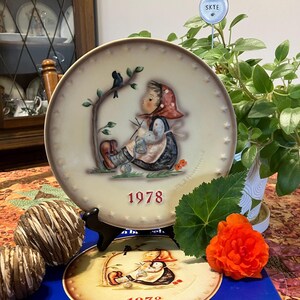May include: A vintage 1978 Goebel plate featuring a girl in a red and white dress knitting with a cat and birds in the background. The plate is on a blue and white box with the Goebel logo.