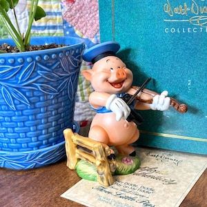 May include: A figurine of a smiling pig playing a violin, wearing a blue hat and vest, and white gloves. The figurine is part of the Walt Disney Collection and is accompanied by a certificate of authenticity. A blue ceramic pot with a plant is in the background.