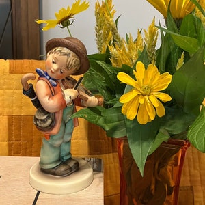 Peut inclure: Une figurine d'un jeune garçon jouant du violon, vêtu d'une tenue bleu et blanche, debout sur un socle blanc. La figurine est entourée de fleurs jaunes dans un vase.