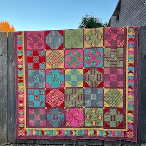 Pode incluir: Um cobertor colorido com um design de patchwork que apresenta várias formas geométricas e padrões em tons de rosa, vermelho, verde, azul e amarelo. O cobertor é enfeitado com uma borda amarela e verde.