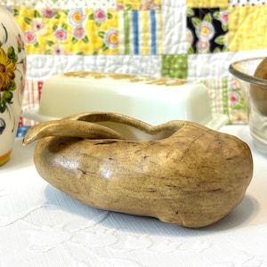 Pode incluir: Um prato de cerâmica em forma de amendoim na cor castanha, com um acabamento liso e brilhante e manchas castanhas escuras. O prato apresenta uma forma alongada e curva com um centro oco e uma pequena pega presa a um dos lados.