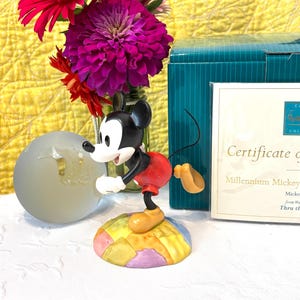Può includere: Una statuetta di Topolino su una base patchwork multicolore, con testa nera, viso bianco, pantaloncini rossi e scarpe gialle. Accanto si trovano una scatola color ottanio e un certificato di autenticità con la scritta Millennium Mickey.