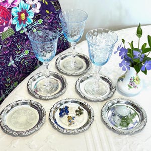 May include: A collection of vintage glassware and serving pieces. The image features three light blue etched wine glasses, each on a silver-colored coaster. Additional coasters and a small vase with flowers are also present, creating an elegant table setting.