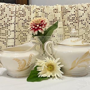 May include: A white porcelain creamer and sugar bowl set with gold accents and a lily of the valley design. The set is on a white crocheted tablecloth.