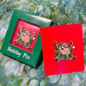May include: A Holiday Pin featuring the Tasmanian Devil character from Looney Tunes, with a green wreath and the year 1997. The pin is displayed on a red card and in a green box with the words "Holiday Pin".